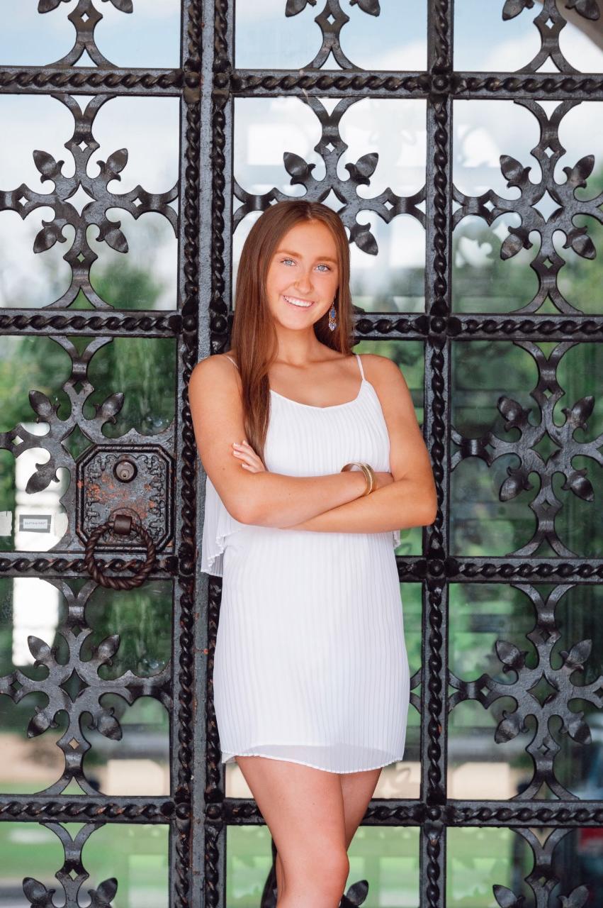 Avery, smiling and wearing a white dress standing in front of a glass and wrought-iron door. 